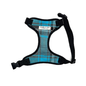 May include: A blue and grey plaid dog harness with black adjustable straps and a buckle fastening. The harness has a label that reads "Barkley & Fetch Canine Outfitters".