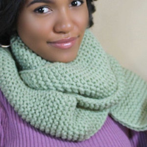 May include: A woman wearing a light green knitted infinity scarf. The scarf is made with a chunky yarn and has a simple stitch pattern.