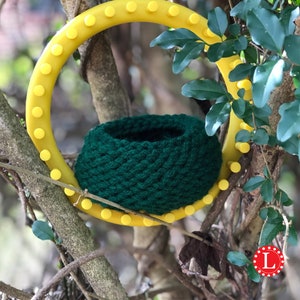 May include: A green knitted bowl sits on a yellow plastic knitting loom with a circular shape. The loom is attached to a tree branch.
