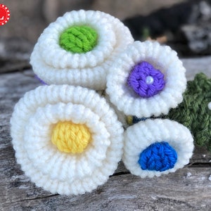 May include: A cluster of crocheted flowers in white, green, yellow, blue, and purple. The flowers are arranged on a piece of weathered wood.