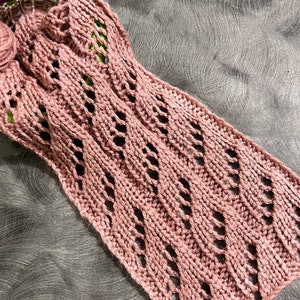 May include: A pink knitted scarf with a lacy pattern. The scarf is lying on a gray textured surface.