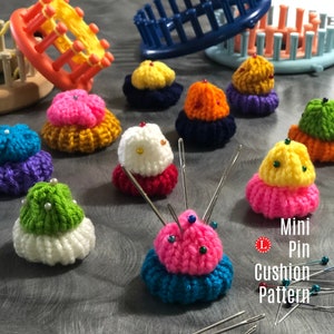 May include: A collection of colourful crocheted pincushions in various shapes and sizes. The pincushions are made with different coloured yarn and have a variety of embellishments, including beads and buttons. The pincushions are arranged on a grey surface with sewing pins scattered around them. The text "Mini Pin Cushion Pattern" is visible in the bottom right corner of the image.