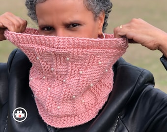 May include: A light pink knitted cowl with a textured pattern and small white pearl accents. The cowl is made of soft yarn and is designed to be worn around the neck for warmth and style. The cowl is a cozy accessory.