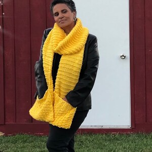 May include: A woman wearing a black jacket and black pants is standing in front of a red barn. She is wearing a bright yellow knitted scarf with pockets.
