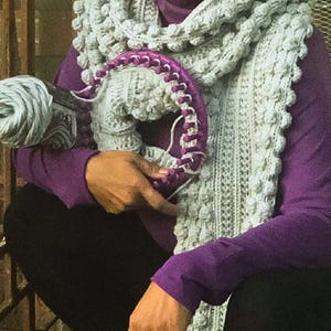 May include: A woman wearing a purple shirt and black trousers is crocheting a grey and white scarf. She is holding a skein of light blue yarn and a purple crochet hook.