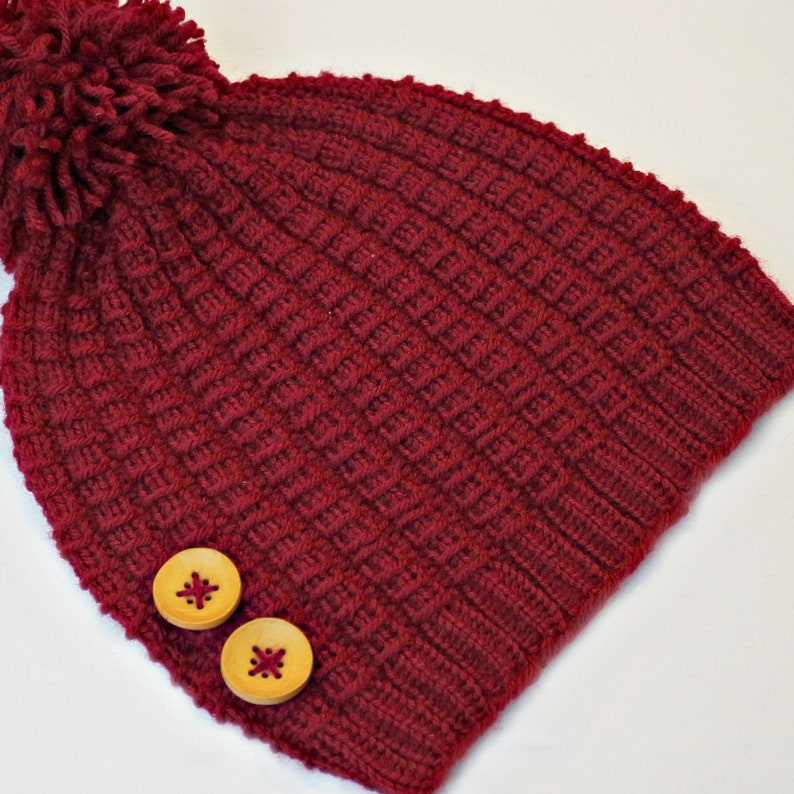 May include: A burgundy knitted beanie with a pom pom on top and two wooden buttons. The beanie has a ribbed texture.