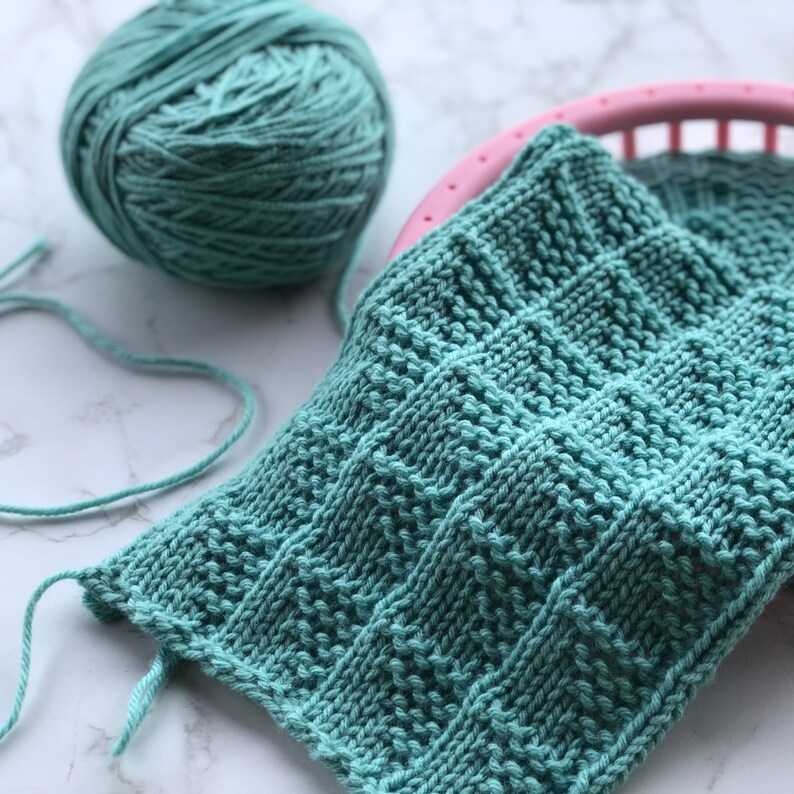 May include: A close-up of a teal knitted square with a textured pattern, partially on a pink loom. A ball of teal yarn sits to the left. The knitted piece has a detailed stitch design, showcasing the texture and craftsmanship of the handmade item.
