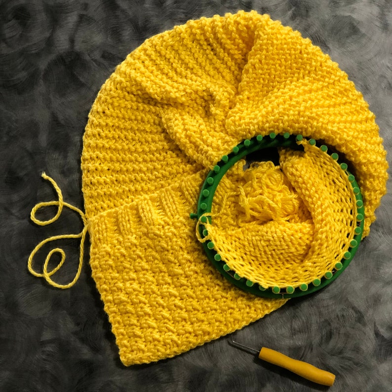 May include: A yellow knitted hat with a green circular knitting loom. The hat is made with a simple stitch pattern and is partially wrapped around the loom.