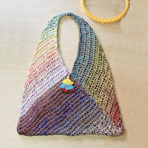 May include: A handmade, open-weave tote bag in a rainbow of colors, including red, orange, yellow, green, blue, and purple. The bag features a long strap and a decorative tassel with yellow, blue, and red accents. A yellow knitting loom is in the background.