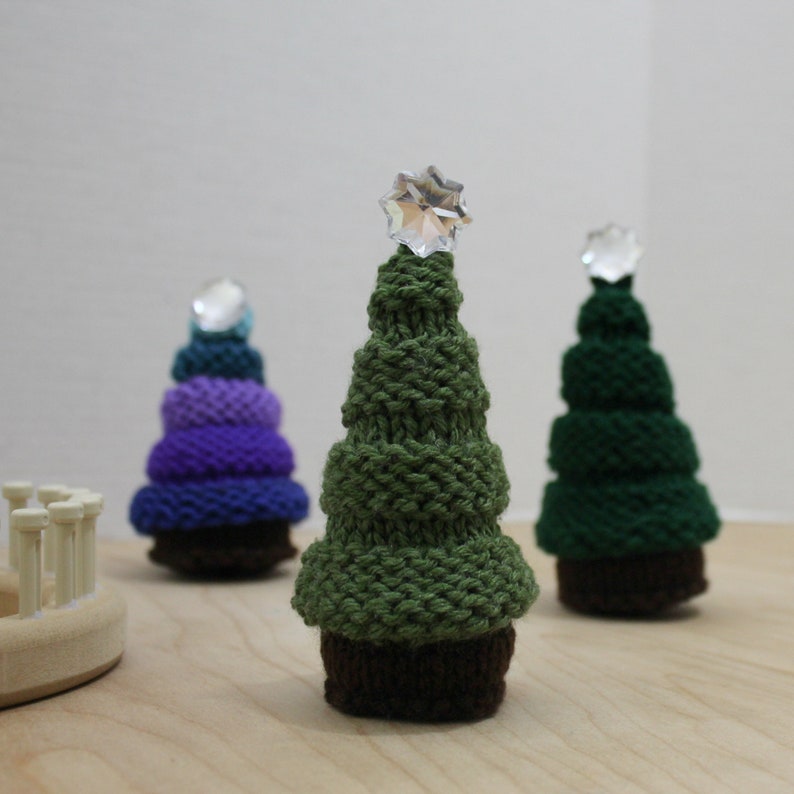 Puede incluir: Tres &aacute;rboles de Navidad tejidos, uno verde, uno morado y azul, y uno azul verdoso. Cada &aacute;rbol tiene una estrella de cristal en la parte superior.