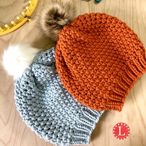 Puede incluir: Dos gorros de punto hechos a mano con pompones de piel sint&eacute;tica. Un gorro es azul claro con un pomp&oacute;n blanco, y el otro es naranja con un pomp&oacute;n marr&oacute;n. Los gorros se muestran sobre una superficie de madera. Un telar amarillo es visible en el fondo.