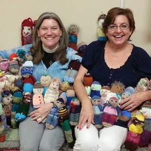 May include: Two women are sitting on a couch surrounded by a large collection of knitted dolls. The dolls are in various colors and patterns, and some have different facial expressions. The women are smiling and appear to be enjoying themselves.