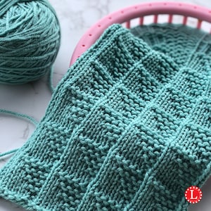 Può includere: Primo piano di un quadrato lavorato a maglia turchese con un motivo strutturato, accanto a un gomitolo di filato turchese e un telaio per maglieria rosa. Il pezzo lavorato a maglia ha un design geometrico. Il filato e il pezzo lavorato a maglia sono su una superficie bianca.