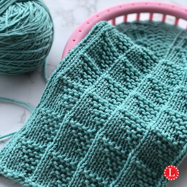 May include: A close-up of a teal knitted square with a textured pattern, next to a ball of teal yarn and a pink knitting loom. The knitted piece has a geometric design. The yarn and knitted piece are on a white surface.