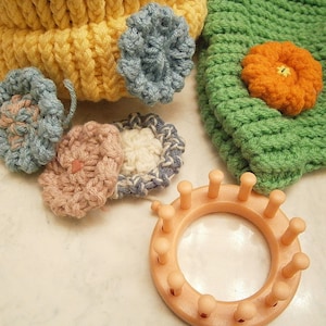 May include: A close-up of a crocheted hat with a variety of colorful crocheted flowers attached. A plastic knitting loom is also in the image.