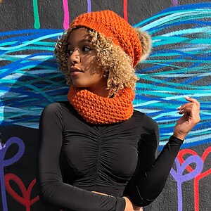 Puede incluir: Un gorro naranja tejido con un pomp&oacute;n de piel sint&eacute;tica y un calentador de cuello a juego. El gorro y el calentador de cuello est&aacute;n hechos de un material de punto grueso. La persona lleva una camisa negra de manga larga.