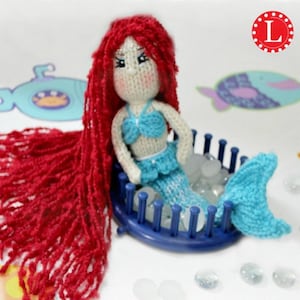 May include: A handmade mermaid doll with red yarn hair, a blue crocheted top and tail, and white skin. The doll is seated in a blue loom. The doll has a sweet face with rosy cheeks and is surrounded by clear, round beads.