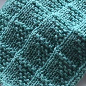 May include: Close-up of a teal knitted fabric with a textured, geometric pattern. The knitwear features a series of diagonal and vertical lines, creating a raised, tactile surface. The yarn appears soft and the color is a muted, seafoam green.
