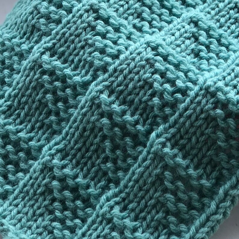 May include: Close-up of a teal knitted fabric with a textured, geometric pattern. The knitwear features a series of diagonal and vertical lines, creating a raised, tactile surface. The yarn appears soft and the color is a muted, seafoam green.
