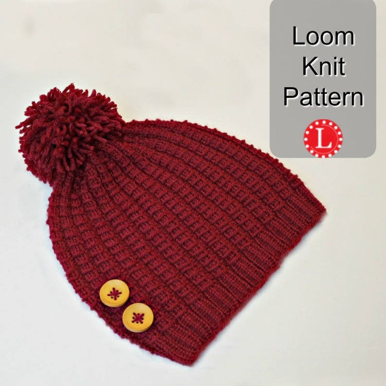 May include: A burgundy knitted beanie with a fluffy pom pom on top and two wooden buttons on the side. The beanie is made with a loom knitting technique. The text "Loom Knit Pattern" is visible in the image.