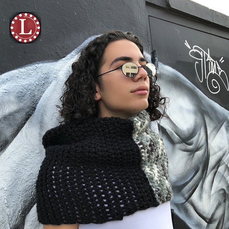 May include: A black and gray knitted cowl and scarf set. The cowl is a solid black with a textured, open weave design. The scarf is a mix of gray, white, and brown. The set is worn over a white shirt.