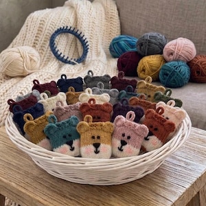 May include: A white woven basket filled with numerous small, hand-knitted bear-shaped pouches in various colours including teal, mustard yellow, pink, and brown. Each pouch features a stitched face with eyes, a nose, and pink cheeks.