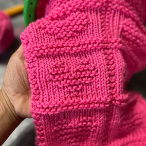 Könnte beinhalten: Nahaufnahme eines rosa gestrickten Stoffes mit einem strukturierten Muster. Der Stoff ist weich und hat ein zartes, weibliches Gefühl.