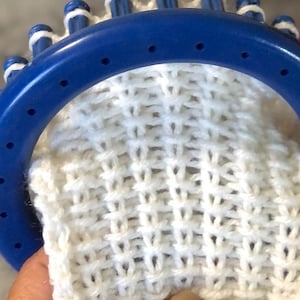 May include: A close-up of a white knitted fabric being worked on a blue circular knitting loom. The fabric has a textured, open-weave pattern.
