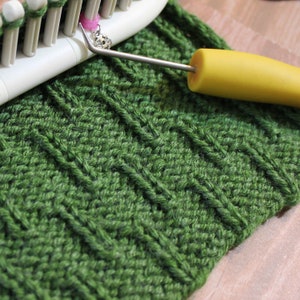 May include: Close-up of a green knitted fabric with a textured pattern. A yellow handled crochet hook is lying on top of the fabric.