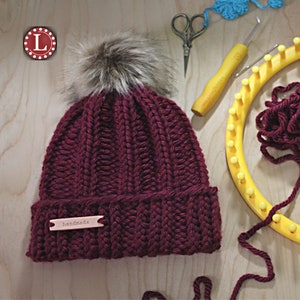May include: A burgundy knitted beanie with a faux fur pom pom on top. The beanie has a brown leather tag that says "handmade".