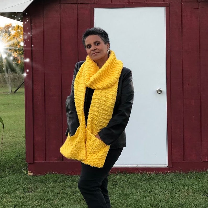 May include: A woman wearing a black jacket and black pants is standing in front of a red barn. She is wearing a bright yellow knitted scarf with pockets.