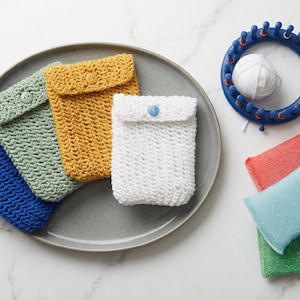 Peut inclure: Trois pochettes rectangulaires au crochet en bleu, vert sauge et jaune moutarde, chacune avec un bouton de fermeture. Une pochette blanche au crochet avec un bouton bleu, un métier à tricoter circulaire bleu avec de la laine et trois éponges.