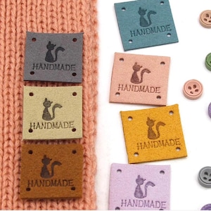 May include: Six different colored leather patches with a cat silhouette and the word "HANDMADE" printed on each. The patches are arranged in a row on a pink knitted surface.