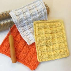 Peut inclure: Trois carr&eacute;s de d&eacute;barbouillettes tricot&eacute;es &agrave; la main en blanc, orange et jaune, avec un motif carr&eacute; textur&eacute; en relief. Un m&eacute;tier &agrave; tisser en bois et un crochet avec un manche en bois sont &eacute;galement visibles, sugg&eacute;rant un projet artisanal.