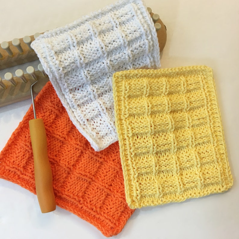 Peut inclure: Trois carr&eacute;s de d&eacute;barbouillettes tricot&eacute;es &agrave; la main en blanc, orange et jaune, avec un motif carr&eacute; textur&eacute; en relief. Un m&eacute;tier &agrave; tisser en bois et un crochet avec un manche en bois sont &eacute;galement visibles, sugg&eacute;rant un projet artisanal.