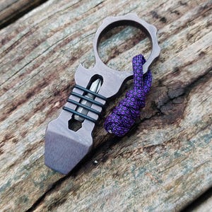 May include: A dark gray metal multi-tool featuring a circular finger hole, a flat pry bar end, and a central slot holding a silver screwdriver bit secured by three black rubber rings. A purple and black paracord lanyard is attached.