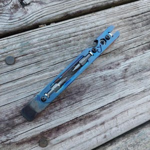 May include: A blue titanium multi-tool pry bar featuring a flat edge, a bottle opener cutout, and two integrated screwdriver bits held in place by black screws. The tool has a metallic finish with visible heat-treated color gradients.