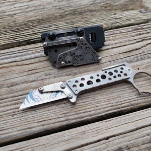 May include: A silver folding knife with a partially serrated blade and a brown handle. The blade has a blue and white design. A black and silver sheath with skull designs is also present.