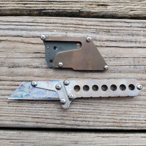 May include: A custom utility knife with a decorative blade and a matching sheath. The blade has a blue and purple pattern, and the handle has a series of holes. The sheath is made of metal and is secured with screws.