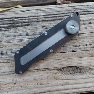 May include: A dark gray utility knife with a silver blade and a circular adjustment wheel. The knife is secured with small silver screws and has a hole for a lanyard. The knife is shown on a weathered wooden surface.