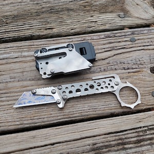 May include: A silver utility knife with a decorative blue blade and a finger ring. The knife has a series of circular cutouts along the handle and is paired with a silver and black tool.