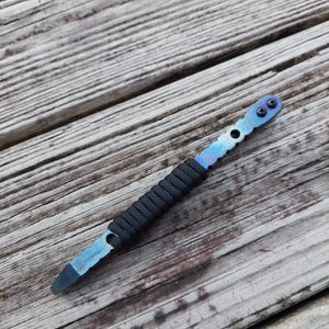 Puede incluir: Una palanca multiherramienta con un acabado degradado en azul y morado anodizado al calor sobre el cuerpo de metal. La sección central está envuelta en paracord negro para mejorar el agarre y el extremo superior incluye dos tornillos negros.