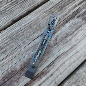 May include: A gray metal multi-tool pry bar featuring a flat edge, a bottle opener, and two integrated screwdriver bits held in place by small black screws. The tool has a dark, iridescent finish and a cutout design along the center.