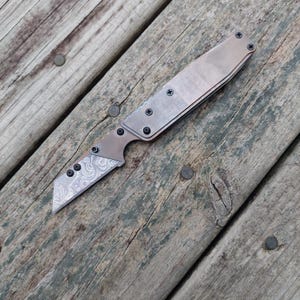 May include: A folding utility knife with a metallic brown handle and a blade featuring a detailed, swirling silver-toned pattern. The handle is secured with multiple small black screws, and the blade has a straight edge with a pointed tip.