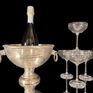 May include: A silver champagne bucket with a bottle of champagne inside, and a pyramid of champagne glasses. The bucket has handles and text that reads "Cuvee de Prestige Champagne De Bauvere". The glasses are clear.