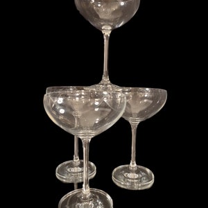 May include: A stack of clear champagne coupe glasses. The glasses have a shallow, wide bowl and a long stem. The glasses are arranged on a black background, creating a visually appealing display. The glassware is ideal for serving champagne or other sparkling wines.