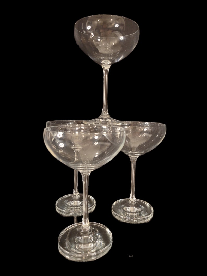 May include: A stack of clear champagne coupe glasses. The glasses have a shallow, wide bowl and a long stem. The glasses are arranged on a black background, creating a visually appealing display. The glassware is ideal for serving champagne or other sparkling wines.