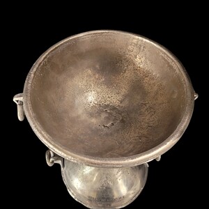 May include: A silver-toned metal bowl with a rounded base and a wide, shallow top. The bowl has two small handles on the sides and a textured surface. The object is set against a black background.