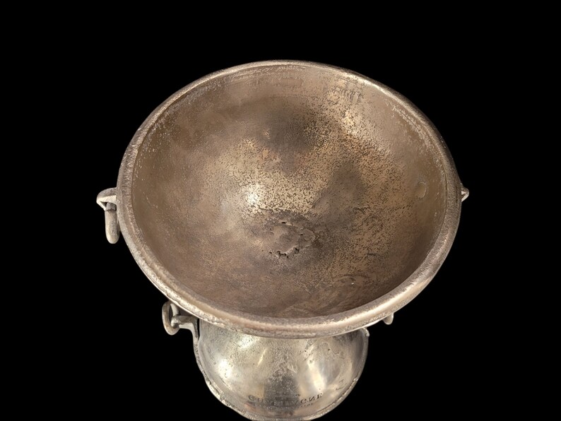 May include: A silver-toned metal bowl with a rounded base and a wide, shallow top. The bowl has two small handles on the sides and a textured surface. The object is set against a black background.