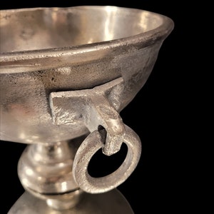 May include: Close-up of a silver-colored metal chalice with a rounded bowl and a ring handle. The chalice sits on a pedestal base, reflecting the light. The background is black.